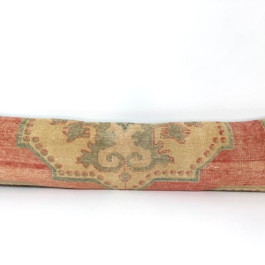 May include: A long, rectangular lumbar pillow featuring a woven textile cover with a faded, distressed appearance. The design displays a central medallion pattern in muted sage green and beige, framed by borders of weathered terracotta red.