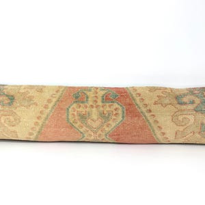 May include: A long, rectangular lumbar pillow featuring a woven textile pattern in muted shades of beige, terracotta orange, and sage green. The design includes a central geometric medallion flanked by intricate floral and vine motifs.