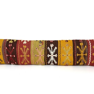 May include: A long, rectangular lumbar pillow featuring a woven textile pattern with vertical stripes in shades of mustard yellow, deep red, and dark brown. The fabric displays repeating geometric motifs in white and light brown throughout the design.