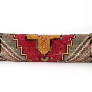 May include: A long, rectangular bolster pillow featuring a woven textile cover with a geometric pattern in shades of deep red, mustard yellow, and gray. The design includes intricate floral-like motifs on the ends and a central medallion.