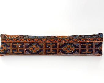 Puede incluir: Un cojín cilíndrico largo y rectangular con una funda de tejido textil sobre una base de color azul marino oscuro. La superficie muestra un patrón geométrico repetitivo en tonos naranja quemado, azul claro y verde, enmarcado por un borde decorativo.