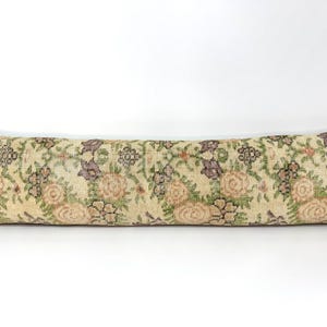 May include: A long, rectangular bolster pillow featuring a woven textile cover with a repeating floral pattern in muted shades of beige, sage green, dusty rose, and dark purple. The fabric has a textured appearance with visible weave details.