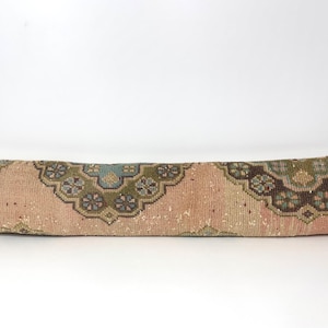 Puede incluir: Un largo cojín rectangular con un aspecto vintage descolorido. El cojín presenta un fondo beige rosado con un patrón repetido de diseños florales en verde azulado, verde y marrón. Probablemente está hecho de un material tejido.