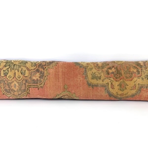 Puede incluir: Un cojín lumbar largo y rectangular con una textura desgastada y descolorida en tonos naranja terracota, verde oliva apagado y gris carbón. La tela muestra un patrón repetitivo de medallones florales ornamentados y simétricos.