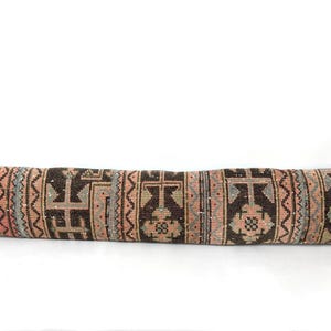 May include: A long, rectangular cushion with a patterned design. The cushion features a repeating geometric pattern in shades of brown, pink, and grey. The cushion is likely used for decorative purposes on a bed or sofa.