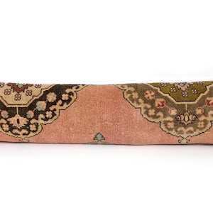 May include: A long, rectangular lumbar pillow featuring a dusty pink base color. The pillow is decorated with two large, symmetrical ornate patterns in shades of dark brown, olive green, and cream, showcasing intricate floral and geometric motifs.