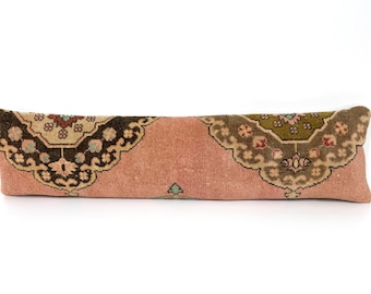 May include: A long, rectangular lumbar pillow featuring a dusty pink base color. The pillow is decorated with two large, symmetrical ornate patterns in shades of dark brown, olive green, and cream, showcasing intricate floral and geometric motifs.