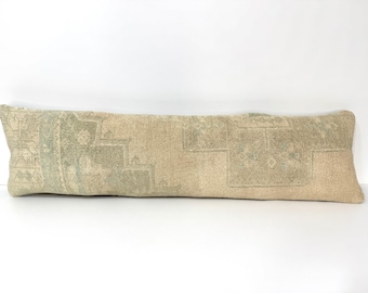 May include: A long, rectangular lumbar pillow featuring a woven textile cover in shades of beige and muted sage green. The fabric displays a faded, intricate geometric pattern with stepped motifs and floral elements across the entire surface.