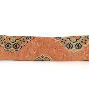 May include: A long, rectangular lumbar pillow featuring a textured, burnt orange base color. The surface is decorated with repeating geometric medallion patterns in shades of teal, navy blue, and cream, creating a symmetrical, woven appearance.