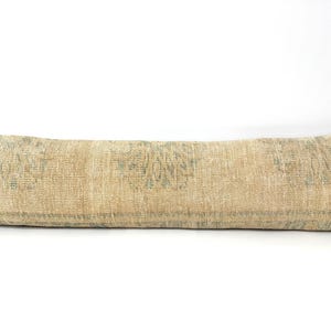 May include: A long, rectangular lumbar pillow featuring a textured, woven fabric in shades of beige and tan. The surface displays a repeating, faded pattern in muted blue-green tones, characterized by intricate, symmetrical floral-like motifs.