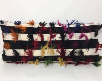 May include: A long rectangular black and white striped pillow with colorful fringe details. The fringe is made of wool and is in various colors including red, blue, green, yellow, and orange.