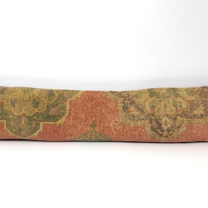 Puede incluir: Un cojín lumbar largo y rectangular con una funda de tejido con un patrón floral intrincado y desteñido en tonos naranja terracota, verde oliva apagado y marrón oscuro, sobre un fondo blanco liso.