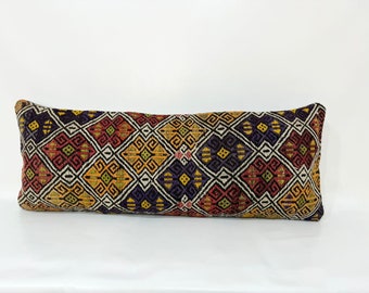 May include: A long, rectangular pillow with a colorful geometric pattern. The pattern features a mix of red, yellow, green, purple, and black colors. The pillow is made of a woven fabric with a textured surface.