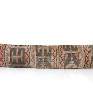 May include: Long, rectangular pillow with a patterned design. The pillow features a mix of colors, including brown, pink, and blue, with geometric shapes and a textured appearance. The pillow is designed for decorative use.