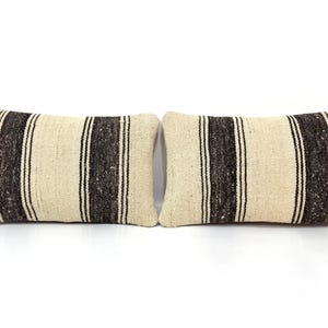 May include: Two rectangular throw cushions featuring a textured, off-white woven fabric base. Each cushion is decorated with two prominent vertical dark brown stripes, which are flanked by thinner black and white pinstripes for contrast.