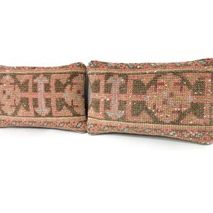 Puede incluir: Dos cojines rectangulares con un patr&oacute;n textil tejido en tonos apagados de rosa empolvado, verde oliva y crema. El dise&ntilde;o consiste en formas geom&eacute;tricas y motivos sim&eacute;tricos que cubren la superficie de ambos cojines decorativos a juego.