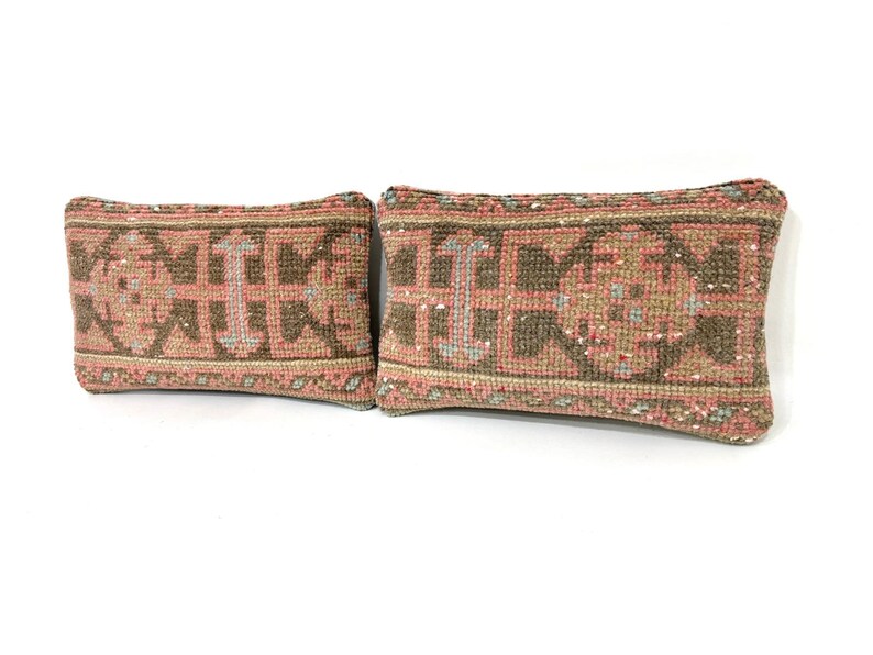 Puede incluir: Dos cojines rectangulares con un patr&oacute;n textil tejido en tonos apagados de rosa empolvado, verde oliva y crema. El dise&ntilde;o consiste en formas geom&eacute;tricas y motivos sim&eacute;tricos que cubren la superficie de ambos cojines decorativos a juego.