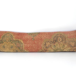 Puede incluir: Un cojín alargado y rectangular de tela texturizada y desgastada en tonos naranja terracota, verde oliva apagado y marrón oscuro. La superficie presenta un patrón floral simétrico y repetitivo a lo largo de toda su extensión.