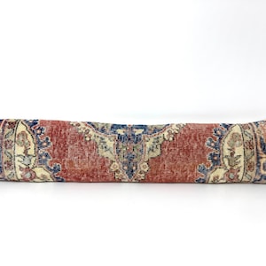 May include: A long, rectangular pillow with a vintage rug design. The pillow features a central section of faded red with intricate blue and cream-colored floral patterns on each end. The pillow is approximately 36 inches long.