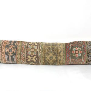 May include: A long, rectangular cushion with a patchwork design. The cushion features a variety of colours, including grey, brown, pink, and green. The design includes geometric and floral patterns. The cushion is likely made of a woven material.