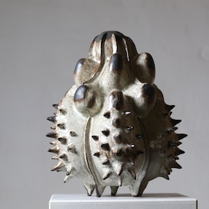 Könnte beinhalten: Eine Keramikskulptur mit cremefarbener Glasur und dunkelbraunen Akzenten. Das Objekt hat einen runden, bauchigen Körper, der mit zahlreichen spitzen Dornen und mehreren glatten, abgerundeten Vorsprüngen im oberen Bereich bedeckt ist.