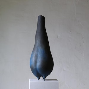 Könnte beinhalten: Eine hohe, tropfenförmige Keramikskulptur mit matter Oberfläche. Die Farbe verläuft von einem tiefen, dunklen Schwarz an der Spitze zu einem leuchtenden, satten Blau an der abgerundeten Basis. Das Objekt steht auf zwei kleinen, spitzen Füßen.