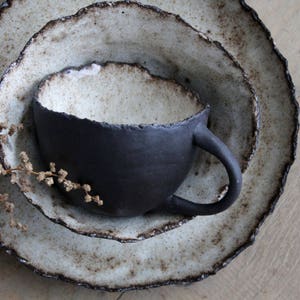 Puede incluir: Un juego de tres platos de cerámica con un esmalte blanco rústico y una taza de cerámica negra con una textura rugosa y desigual. Los platos tienen un borde festoneado y están apilados uno encima del otro.