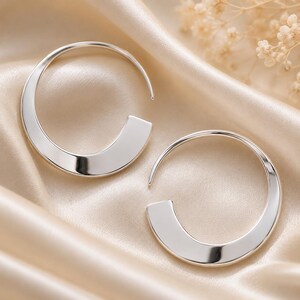 May include: A pair of silver hoop earrings featuring a wide, flat, crescent-shaped bottom section that tapers into a thin, circular wire post. The earrings have a polished, reflective metallic finish and a smooth, curved design.