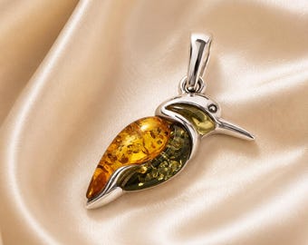 May include: A hummingbird-shaped pendant crafted from polished silver, featuring a body inlaid with translucent golden-yellow and deep forest-green amber. The bird design includes a slender beak, a small eye, and a silver bail at the top for hanging.