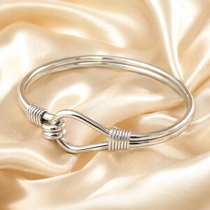 Może przedstawiać: Polerowana bransoletka typu bangle w kolorze srebrnym o konstrukcji z podwójnego drutu, która zbiega się w centralną pętlę przypominającą węzeł. Bransoletka jest zabezpieczona dwoma ozdobnymi sekcjami z owiniętego drutu po obu stronach.