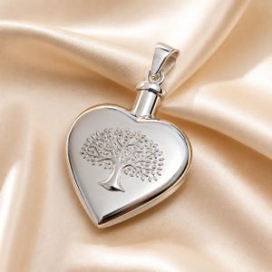 May include: A polished silver-tone heart-shaped pendant featuring a detailed engraved tree design on the front surface. The pendant includes a cylindrical top component with a loop for attachment, showcasing a smooth, reflective metallic finish.