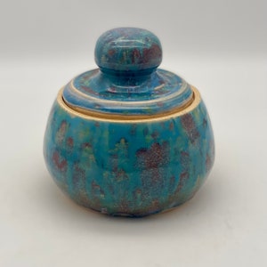 May include: A small ceramic jar with a lid. The jar is a light blue color with brown and green speckles. The lid is a light blue color with brown speckles.