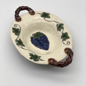 Puede incluir: Un plato de servir de cerámica blanca con dos asas marrones. El plato está decorado con un diseño de vid y un racimo de uvas moradas.