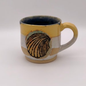 Puede incluir: Una taza pequeña de cerámica amarilla con un diseño de concha blanca y marrón. La taza tiene un borde azul y un asa blanca.
