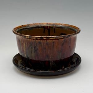 May include: A round ceramic planter with a glossy finish featuring a deep brown and amber drip glaze pattern. The planter sits on a matching circular saucer, both displaying vertical streaks of dark brown and golden tones across the surface.