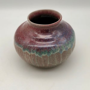 Puede incluir: Un jarrón pequeño y redondo de cerámica con un esmalte rojo-púrpura moteado. El jarrón tiene un esmalte verde claro alrededor del borde inferior.