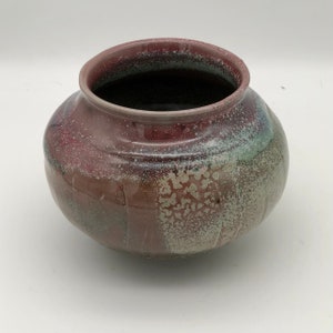 Puede incluir: Un jarrón redondo de cerámica con un esmalte moteado en tonos de rosa, verde y blanco. El jarrón tiene un borde ligeramente acampanado y una base redondeada.