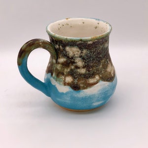 May include: A handmade ceramic mug with a blue, white and brown glaze. The mug has a rounded body and a handle.