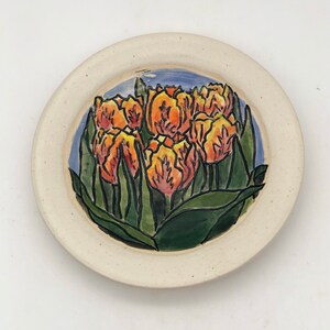May include: A white ceramic plate with a hand-painted design of orange tulips with green leaves and stems. The tulips are arranged in a bouquet on a blue background.