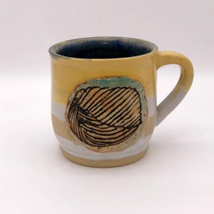 Puede incluir: Una taza de cerámica amarilla con un diseño de hoja blanco y marrón. La taza tiene un glaseado azul claro en el interior.