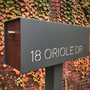 May include: A modern gray mailbox with a wood panel and a silver handle. The mailbox has a white cutout of the address "18 ORIOLE DR".