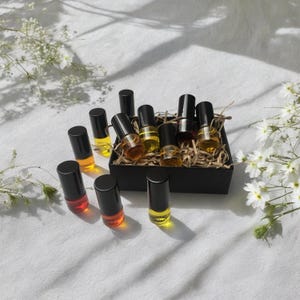 May include: A set of 10 small glass bottles with black roll-on tops filled with a variety of yellow and orange liquids. The bottles are arranged in a small black box lined with brown paper.