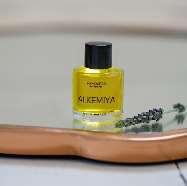 May include: A small glass bottle of yellow liquid with a black cap and label. The label reads "Ink + Ocean Botanicals" and "Alkemiya Parfum Botanique".