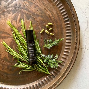 May include: A black roller bottle with a white label that reads "Ink + Ocean Botanicals Aromatherapy Blend Memory Support" on a copper tray with green rosemary and cardamom pods.