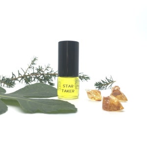 Puede incluir: Una pequeña botella de vidrio transparente con una tapa negra y líquido amarillo en su interior. La botella está etiquetada con "STAR TAKER". La botella está sobre una superficie blanca con hojas verdes y piedras ámbar.
