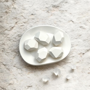 以下が含まれることがあります： 白い楕円形の皿の上に、岩のような白い幾何学的な形が5つ並んでいます。皿は薄い灰色のざらざらした表面の上に置かれています。