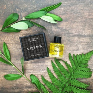 May include: A small bottle of perfume with a black label that says "Alkemiya Ink + Ocean Botanical" and a black box with the same label. The bottle and box are on a wooden surface with green leaves.