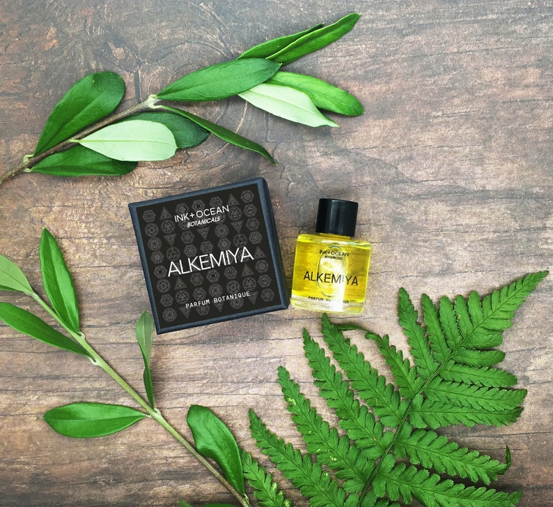 May include: A small bottle of perfume with a black label that says "Alkemiya Ink + Ocean Botanical" and a black box with the same label. The bottle and box are on a wooden surface with green leaves.