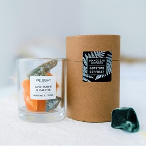 Puede incluir: Un frasco de vidrio lleno de gemas naranjas y verdes, etiquetado con "INK+OCEAN BOTANICALS AVENTURINE & CALCITE GEMSTONE DIFFUSER". Un tubo de cartón marrón con una etiqueta que dice "INK+OCEAN BOTANICALS GEMSTONE DIFFUSER" está al lado del frasco. Una sola gema verde está sobre la mesa.