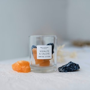 May include: A glass jar filled with blue sodalite and orange calcite gemstones. The jar has a label that reads "Ink + Ocean Botanicals, Sodalite & Calcite, Gemstone Diffuser".